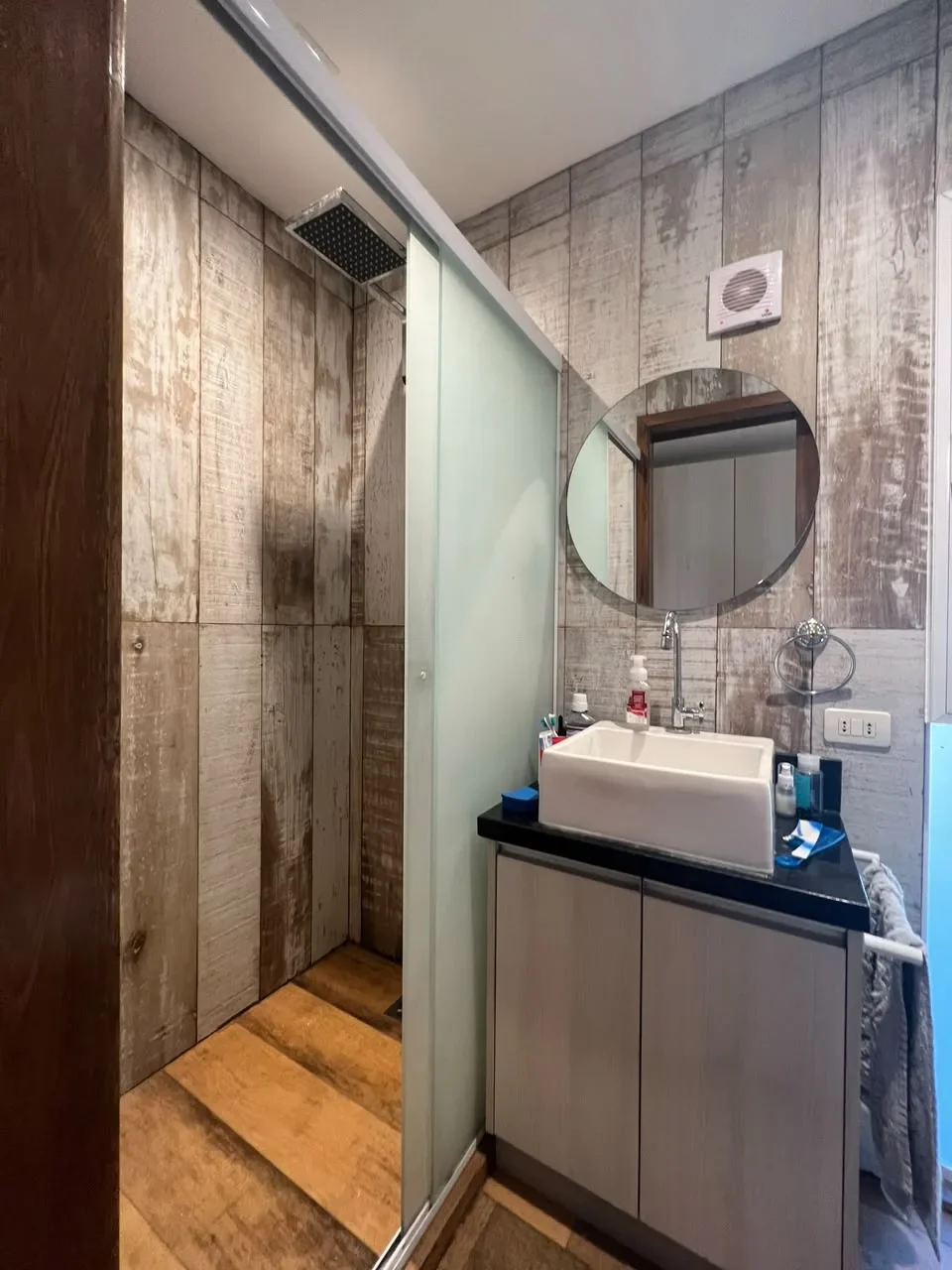 Bano de casa de contenedor con paredes de madera rustica envejecida, box de ducha con puerta esmerilada, espejo redondo y bacha de apoyo sobre granito negro.