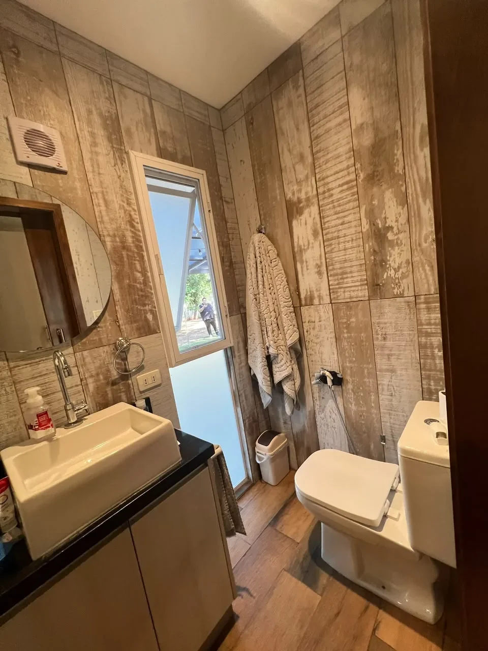 Bano de casa de contenedor con paredes de madera rustica envejecida, espejo redondo, bacha de apoyo sobre mesada de granito negro y ventana al jardin.
