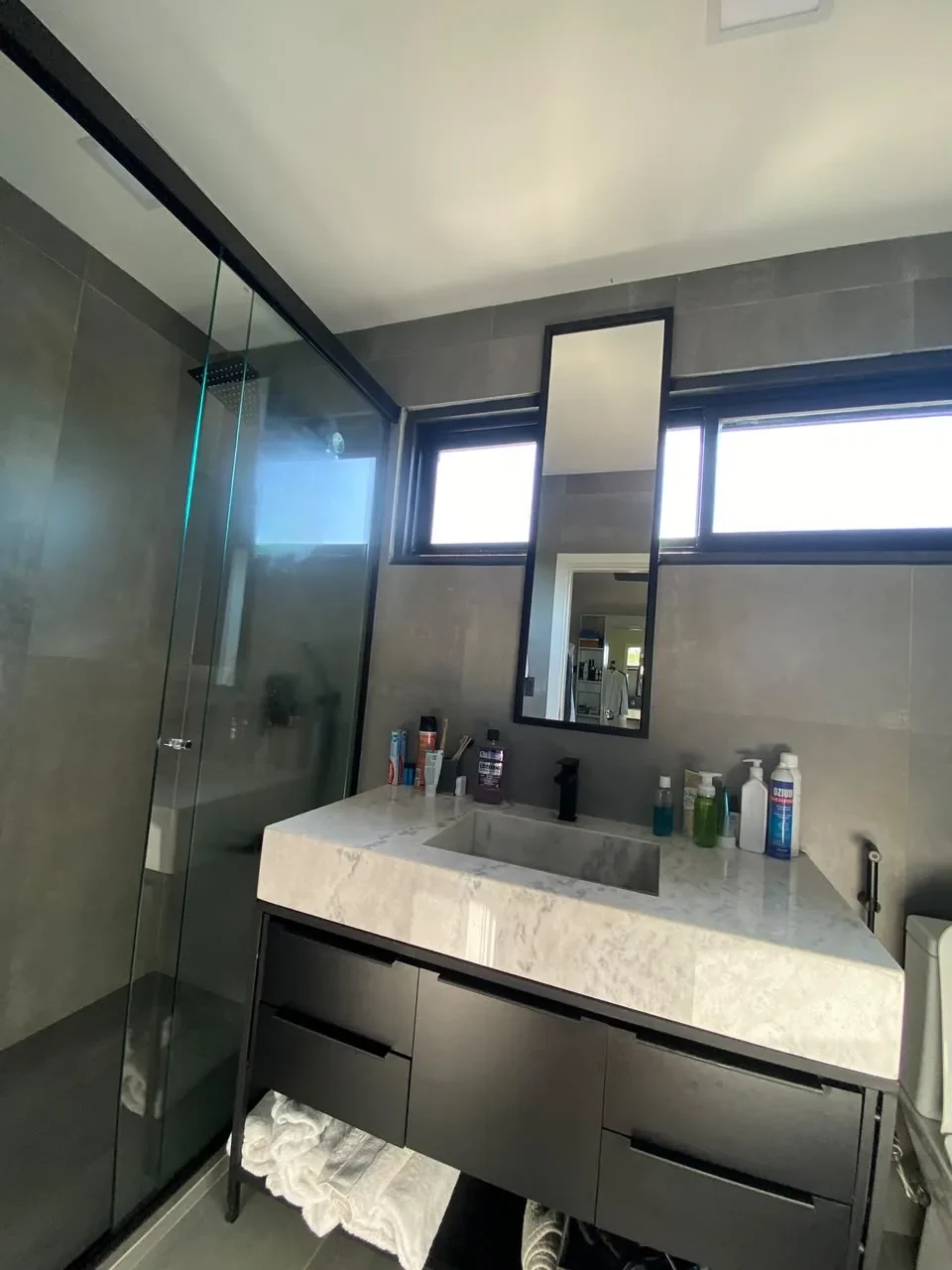 Bano de lujo en casa de contenedor con porcelanato gris, box de ducha de vidrio, vanitory flotante con mesada de marmol y espejo de marco negro.