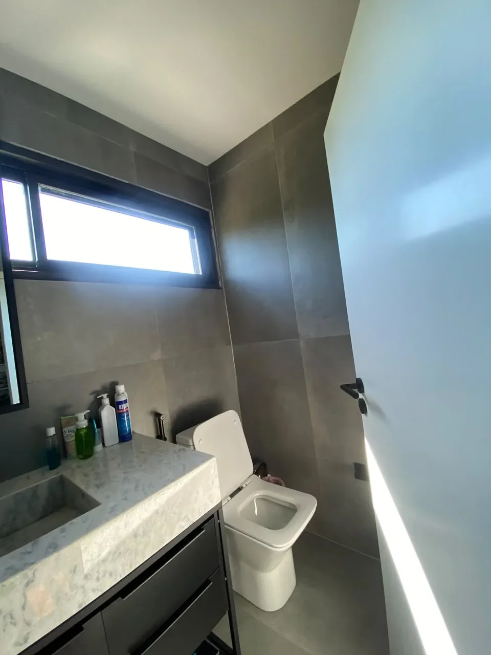 Bano de casa de contenedor con porcelanato gris, vanitory oscuro con mesada de marmol, inodoro y ventana alta esmerilada.
