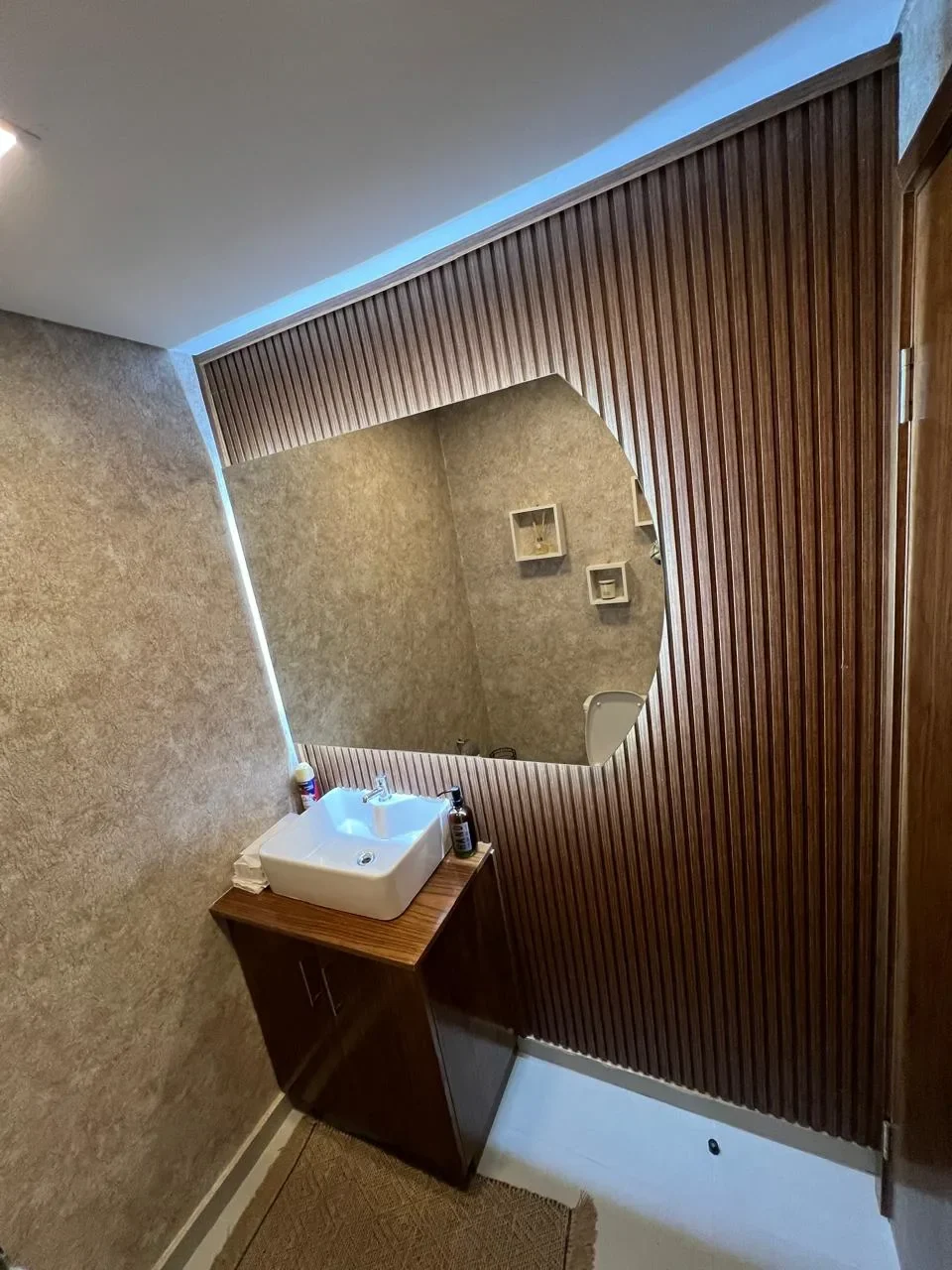 Bano de casa de contenedor con pared de lambri de madera, espejo irregular retroiluminado y bacha de apoyo sobre vanitory de madera.