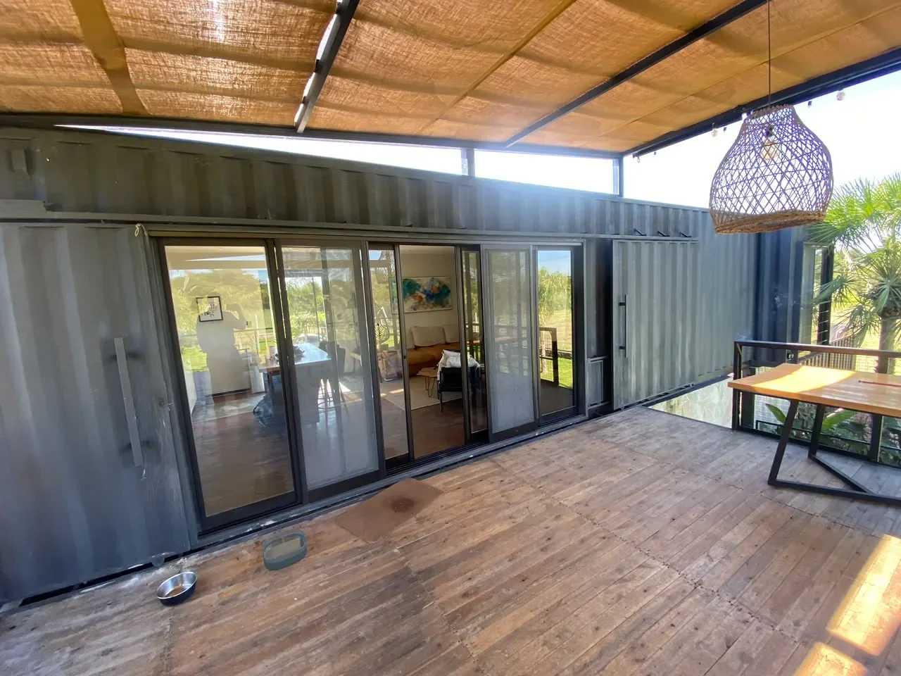 Deck de madera con pergola de una casa de contenedor gris-azulada, puertas corredizas de vidrio, comedor y colgante tejido, vista al jardin.