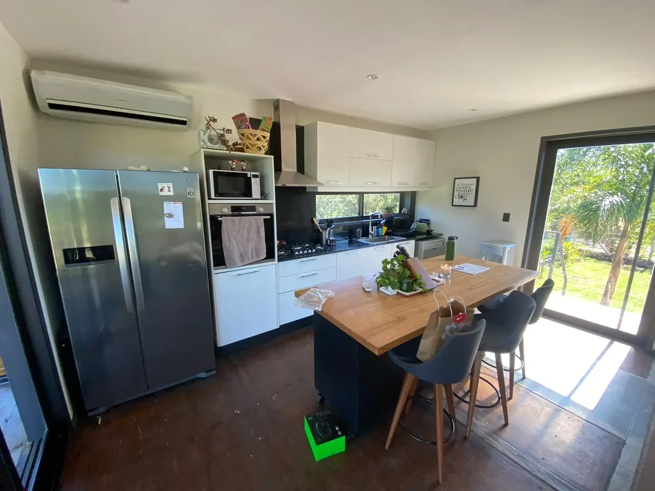 Cocina de casa de contenedor con muebles blancos, mesada negra, isla de madera con banquetas y heladera side-by-side, abierta al jardin por ventanales.