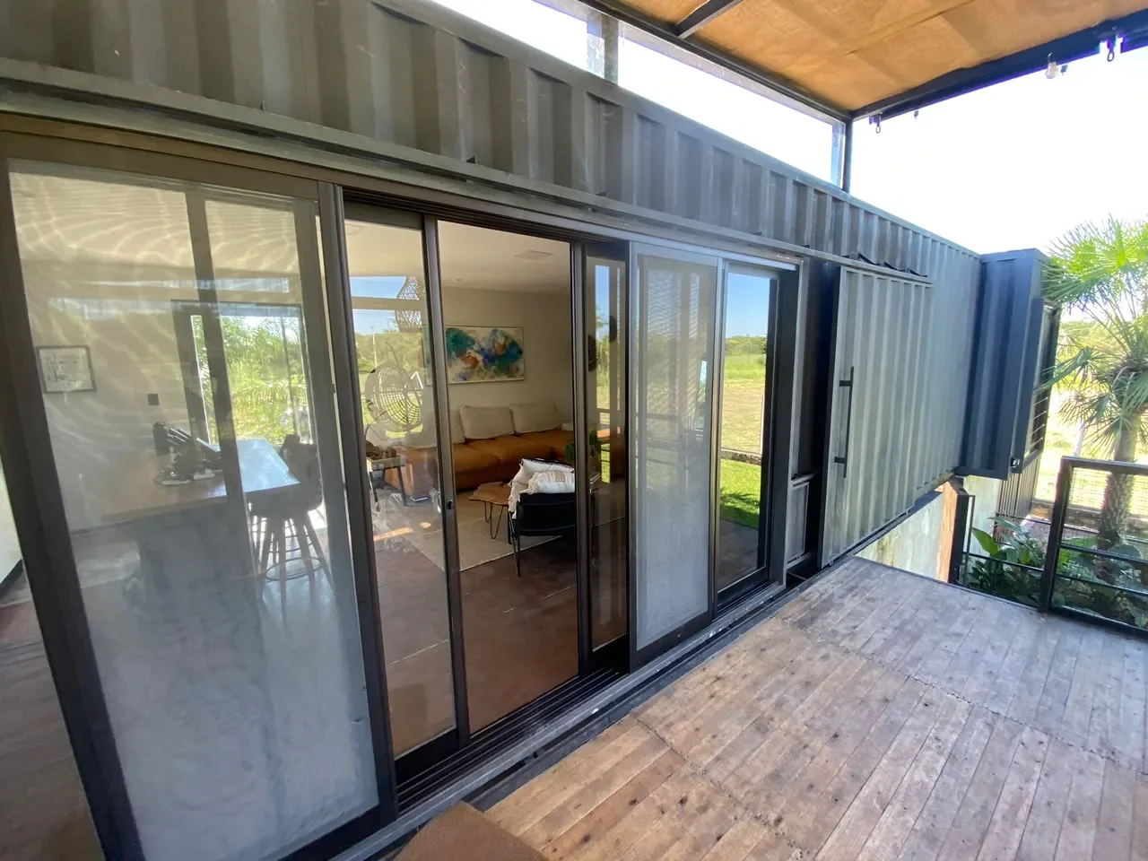 Deck de madera de casa de contenedor oscuro con pergola de cana y puertas corredizas de vidrio que muestran el living, con vista abierta al campo.