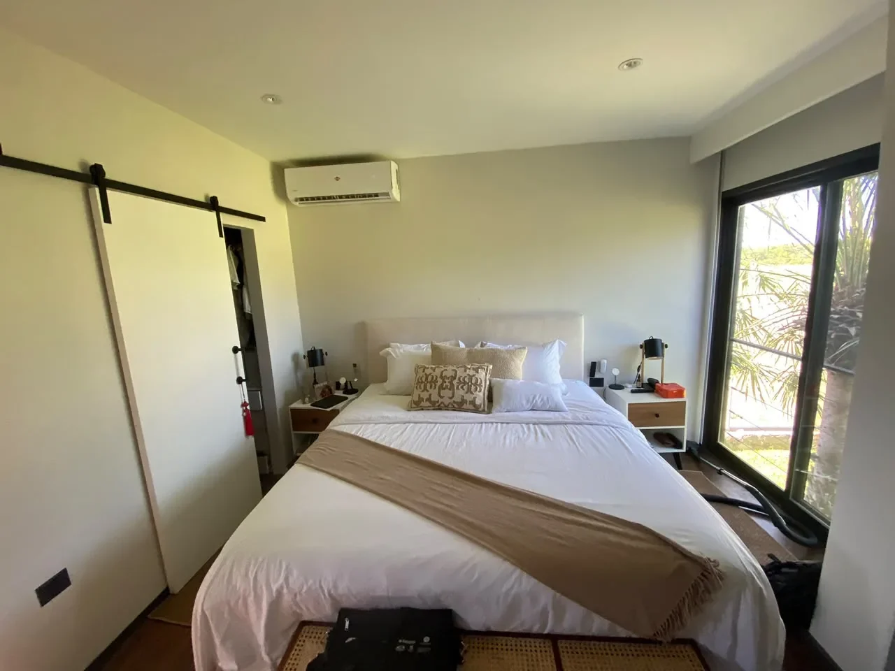 Dormitorio de casa de contenedor con cabecera tapizada, placar de puerta corrediza tipo granero y pared de vidrio hacia el jardin, piso de madera.