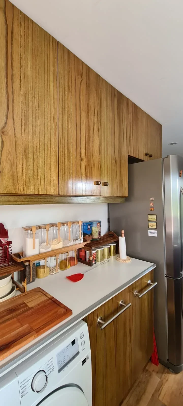 Cocina de casa de contenedor con muebles de enchapado de madera, mesada blanca, lavarropas integrado y heladera, estilo hogareno.
