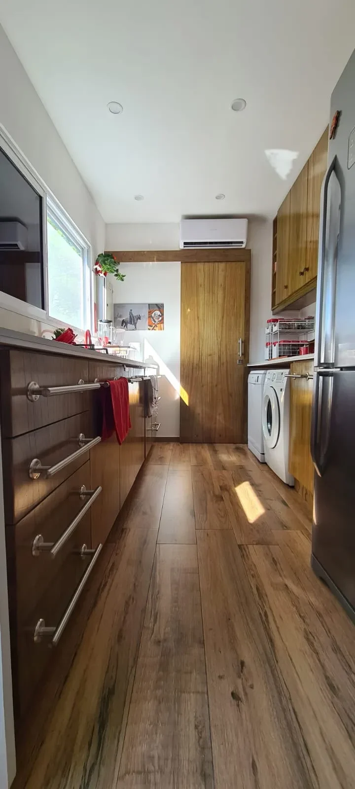 Cocina-lavanderia de casa de contenedor con muebles de madera, mesada blanca, lavarropas, heladera y cuadro ecuestre, piso de madera.