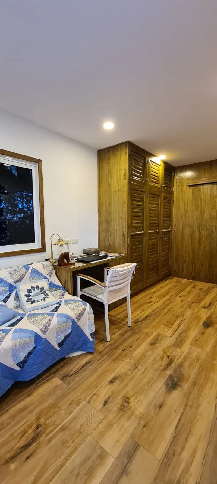 Dormitorio-estudio de casa de contenedor con escritorio, placar de persiana de madera, colcha patchwork azul y ventana a los arboles.
