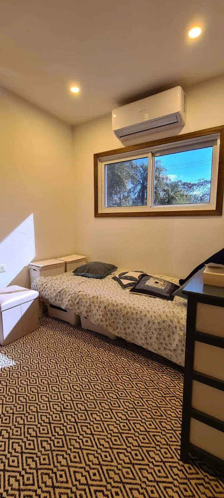 Dormitorio en casa de contenedor con cama individual, alfombra geometrica blanco y negro, ventana con marco de madera y aire acondicionado.