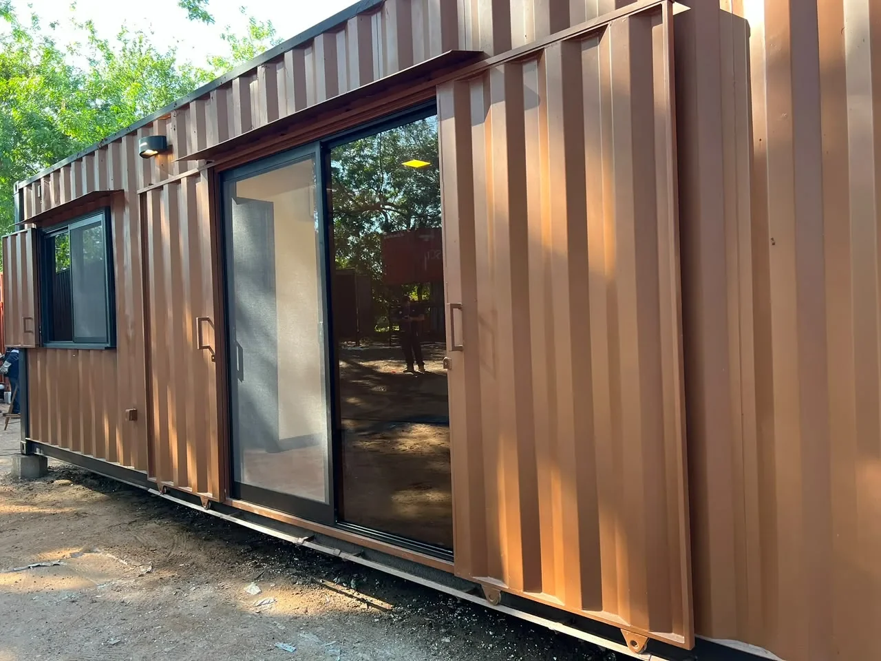 Frente del modulo de contenedor corten marron con puerta corrediza de vidrio y ventanas, entre arboles.
