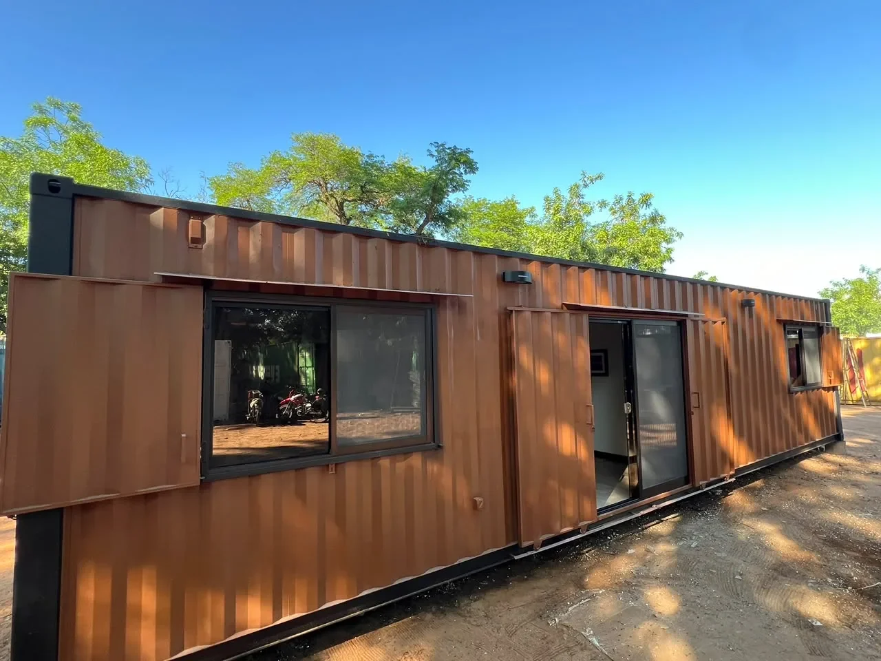 Frente tres cuartos del modulo corten marron con ventana corrediza y puerta abierta mostrando el interior, cielo despejado.