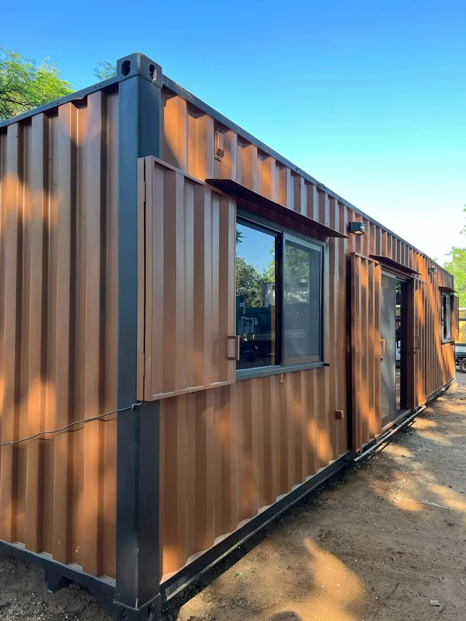 Modulo de contenedor corten marron con postigo rebatible, ventana corrediza y puerta de vidrio, en predio de tierra.
