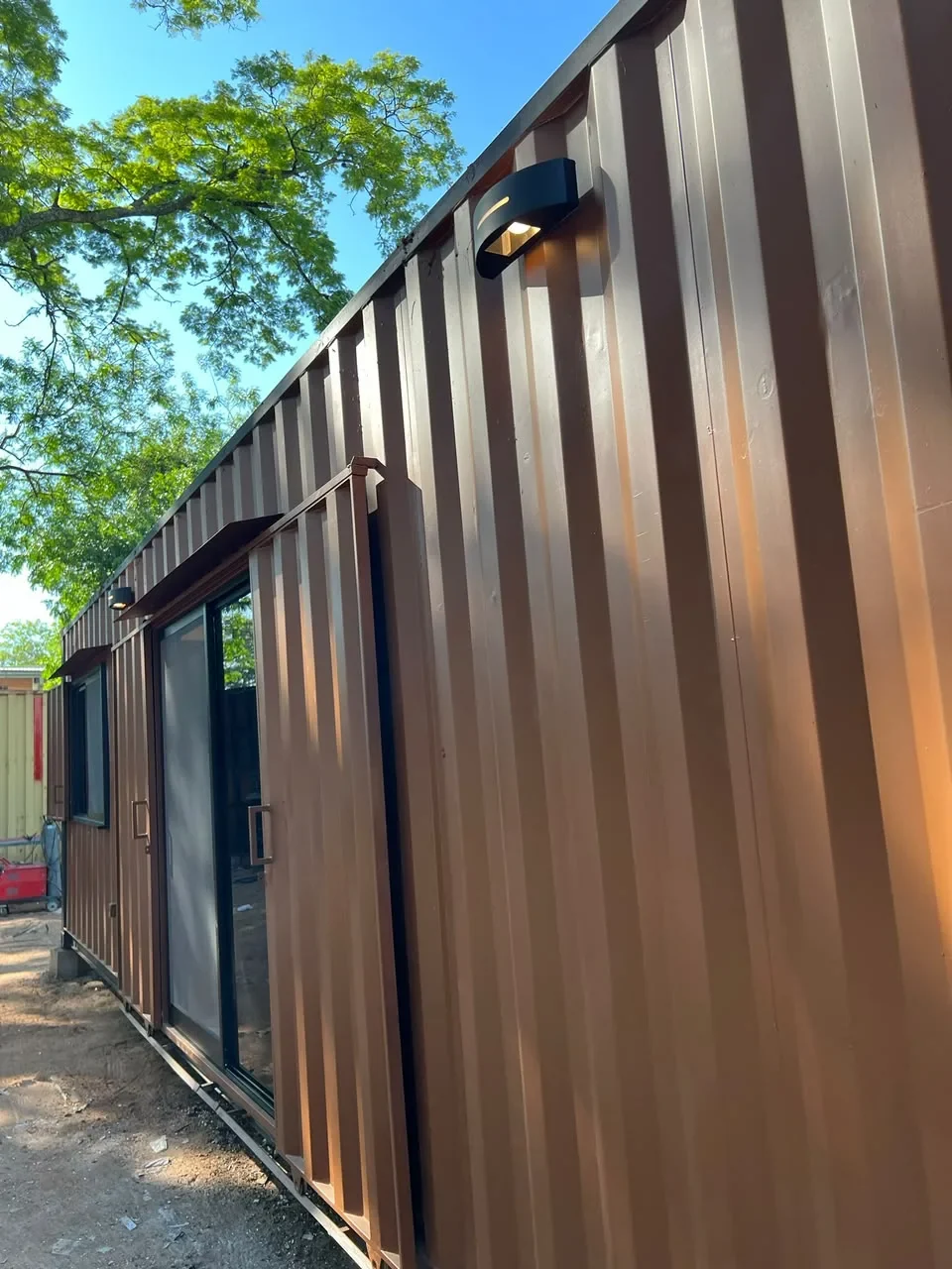 Entrada del modulo corten marron con puerta corrediza de vidrio y aplique de pared negro, entre arboles.